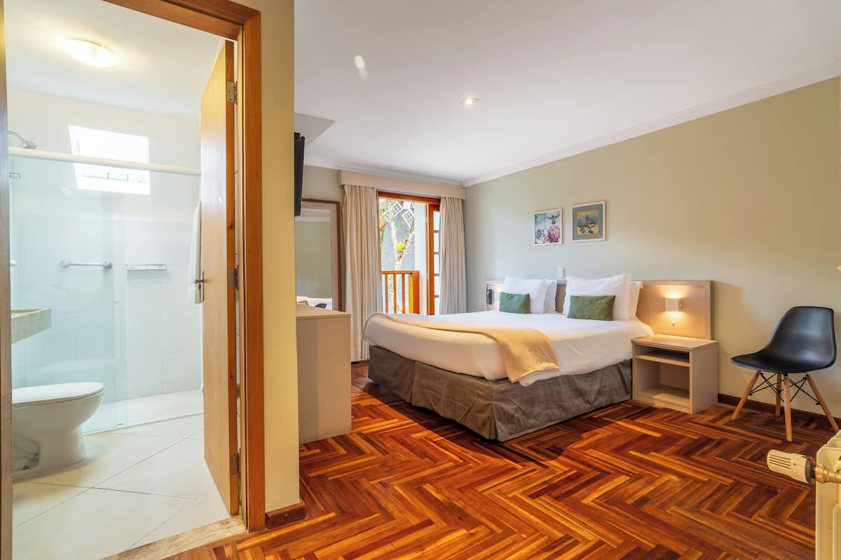 Habitación de hotel con suelo de parquet de madera, cama con ropa blanca, mesita de noche con lámpara, una silla y vistas al exterior a través de una ventana.