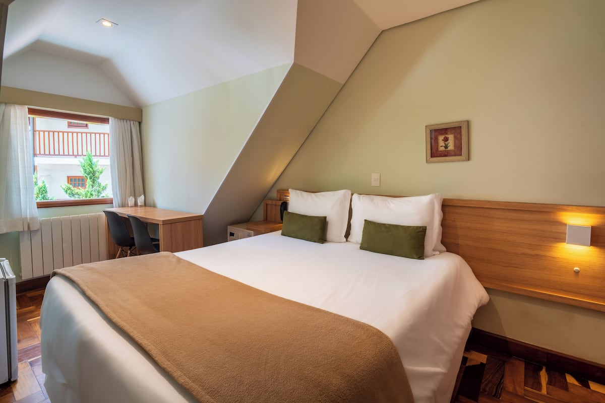 Una habitación de hotel con cama, escritorio, silla, radiador, ventana con cortinas y un cuadro en la pared.