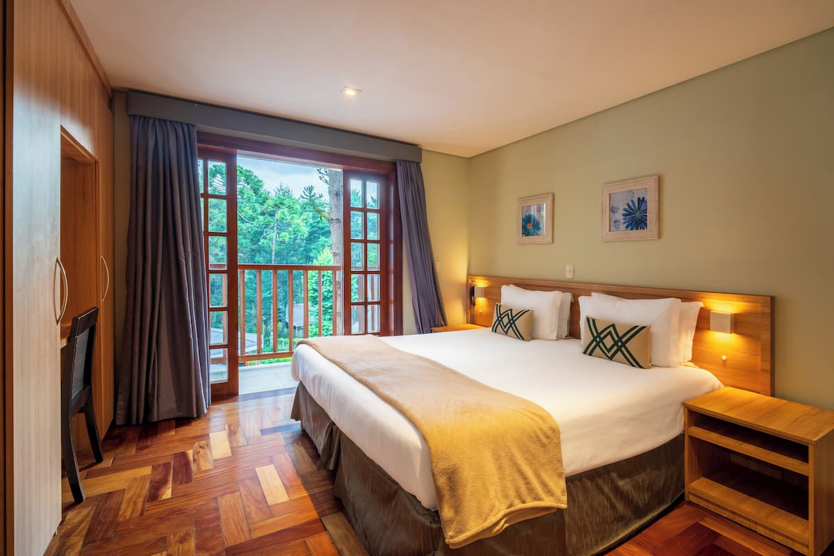 Un dormitorio amplio con una cama grande, piso de madera, un balcón con cortinas y un cuadro en la pared.