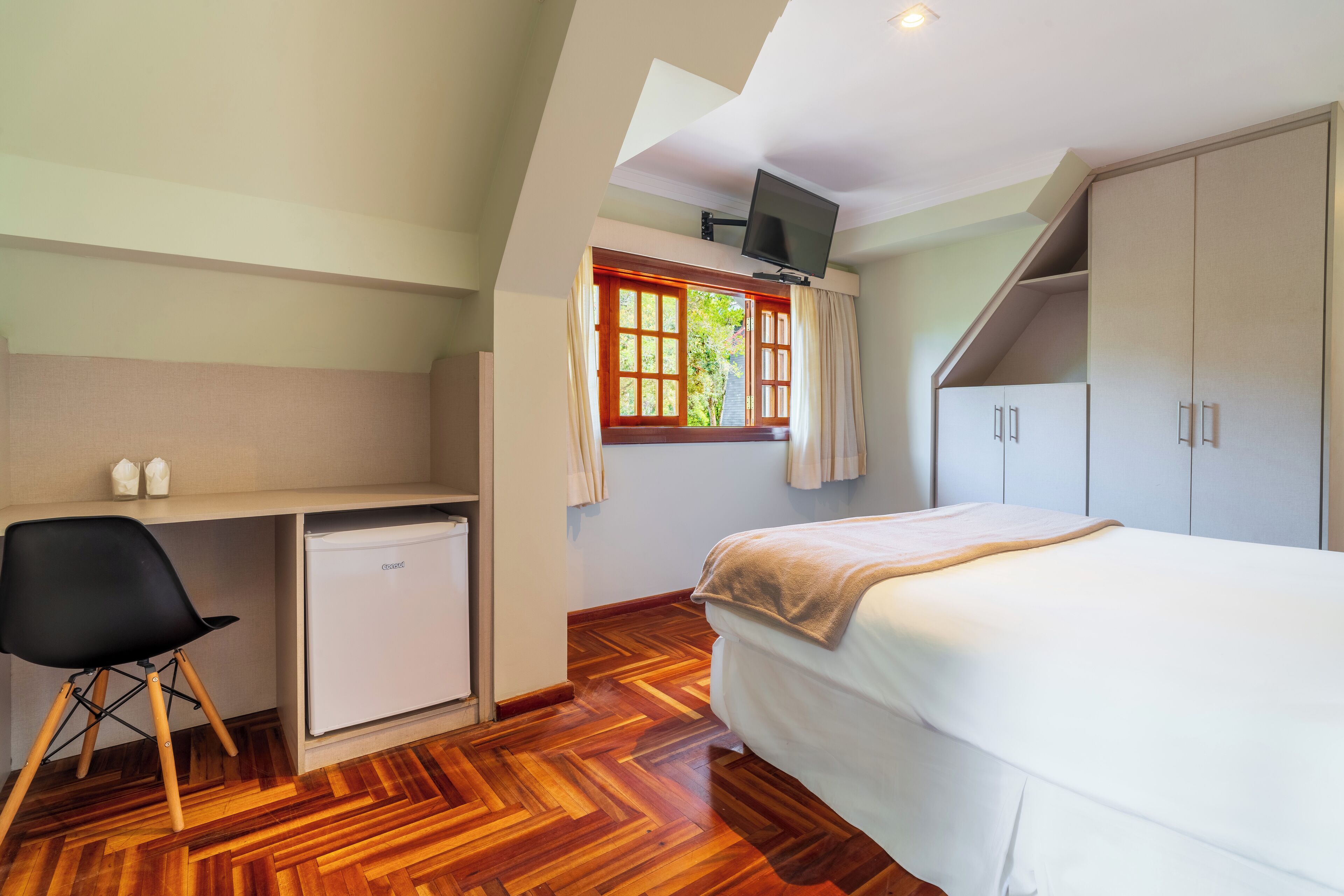 Een slaapkamer met een bed, een bureau met stoel, een kleine koelkast en een aan de muur bevestigde televisie.
