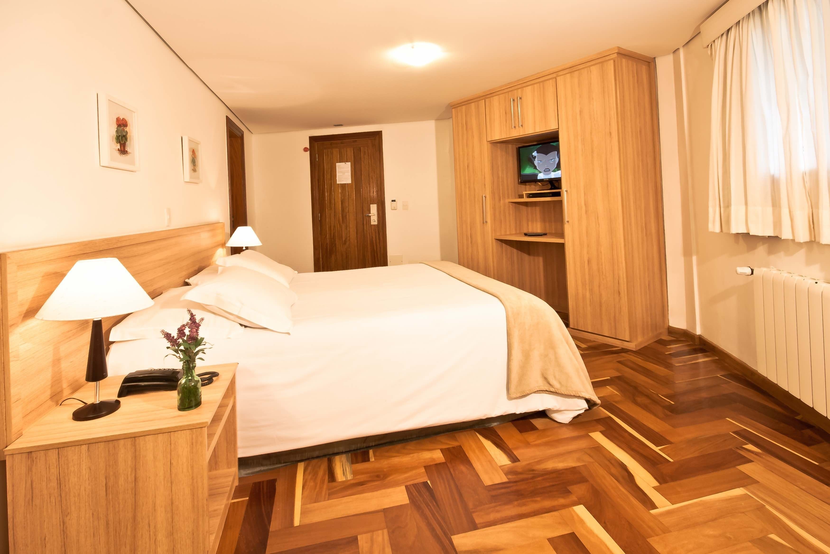 Une chambre d’hôtel avec un lit, une tête de lit en bois, une table de chevet avec une lampe, un petit vase avec des fleurs, une armoire en bois et un téléviseur posé sur une étagère.