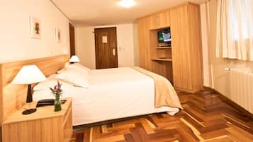 A hotel room with a bed, wooden headboard, bedside table with lamp, small vase with flowers, wooden wardrobe, and a television on a shelf.