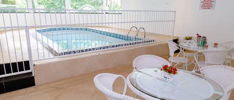 Un área de piscina con una piscina con azulejos, sillas de mimbre blanco y una mesa de cristal con un florero.