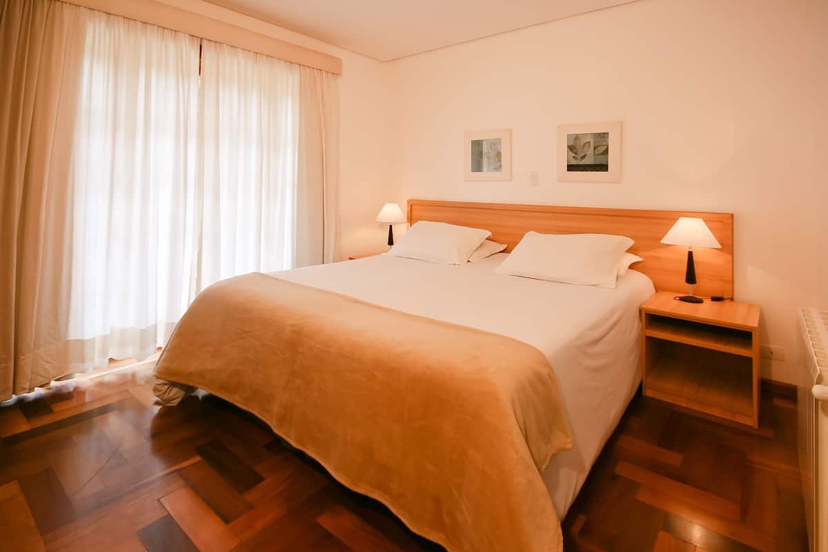 Una cama bien hecha con cabecera de madera, dos lámparas de noche y una ventana con cortinas translúcidas.