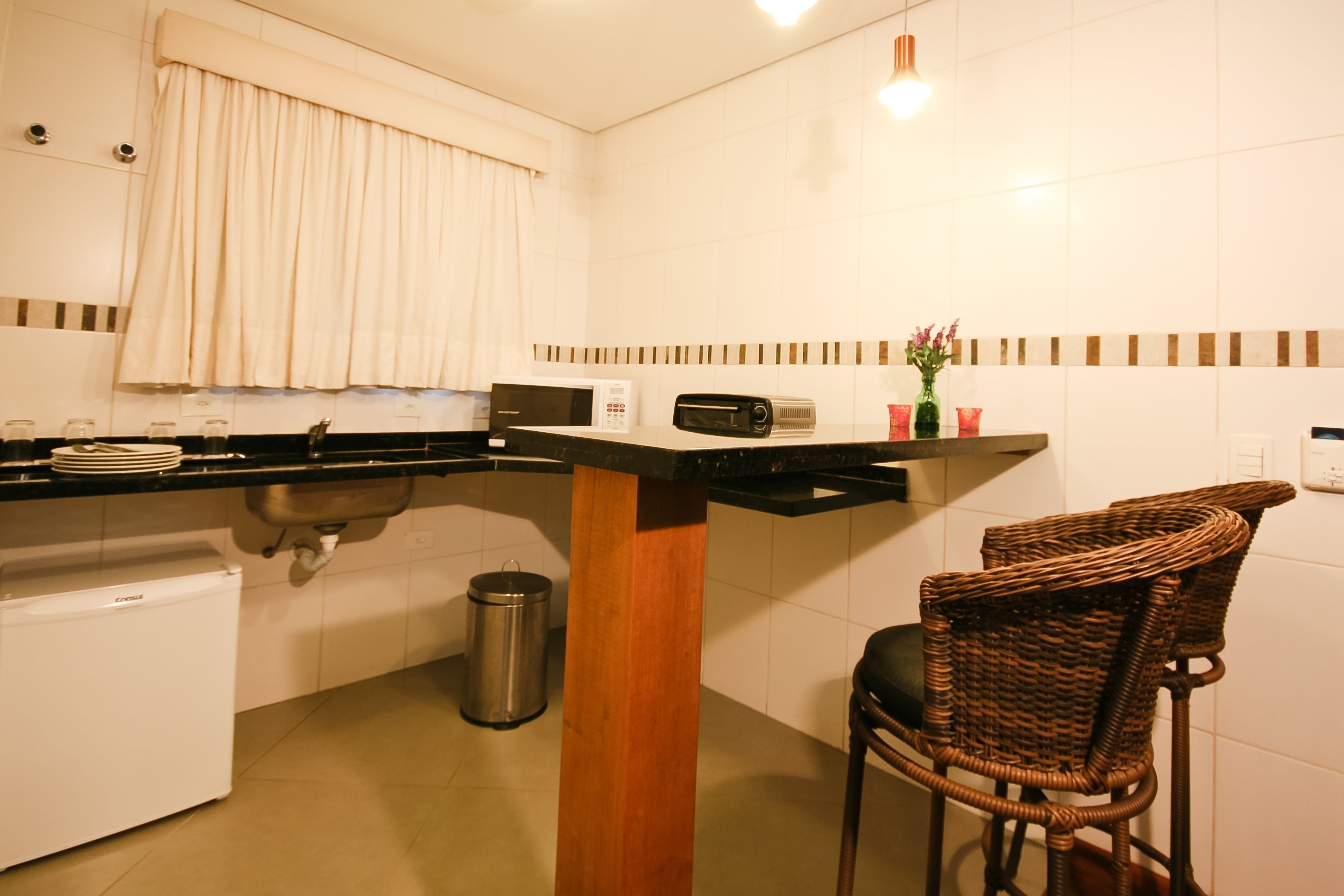 A compact kitchen with a built-in microwave, toaster, and sink. There is a small refrigerator, a trash can, and a wicker chair.