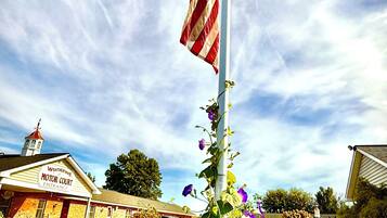 An American flag on a flagpole, a building with a sign reading 'Weatherine Motor Court Entrance,' and purple flowers in the foreground.