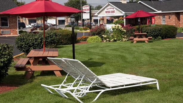 A lawn with a white lounge chair and a red umbrella, a brick building with a sign reading "Weatherlane Motor Court Entrance", and a Coca-Cola sign. - Weathervane Motor Court (Ronks)