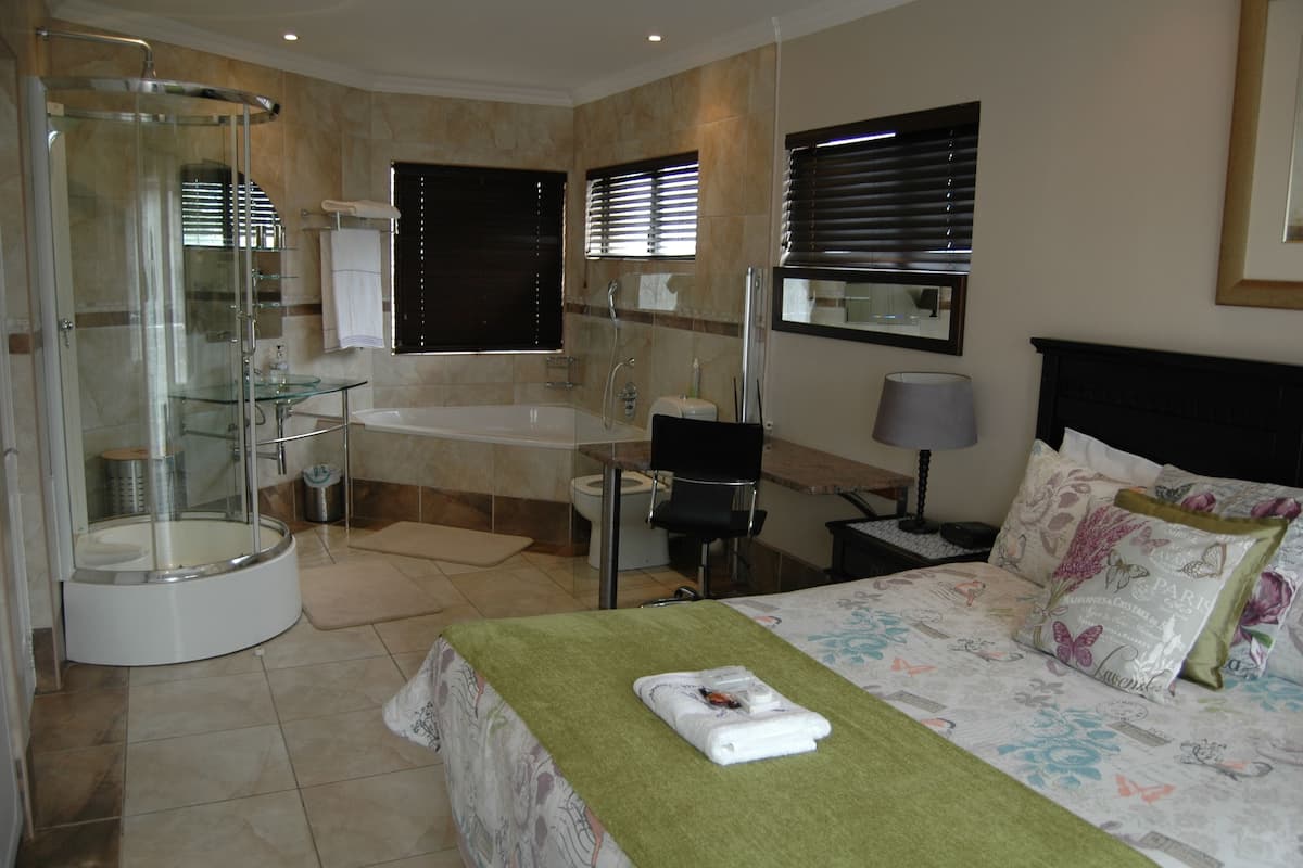 A hotel room with a glass-enclosed shower, a bathtub, a toilet, a sink, a bed with floral bedding, and a bedside table with a lamp.