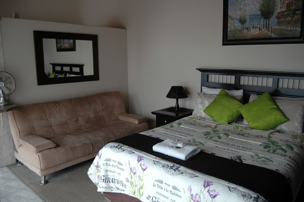 A hotel room with a bed, a sofa, a bedside table with a lamp, and a wall-mounted picture.