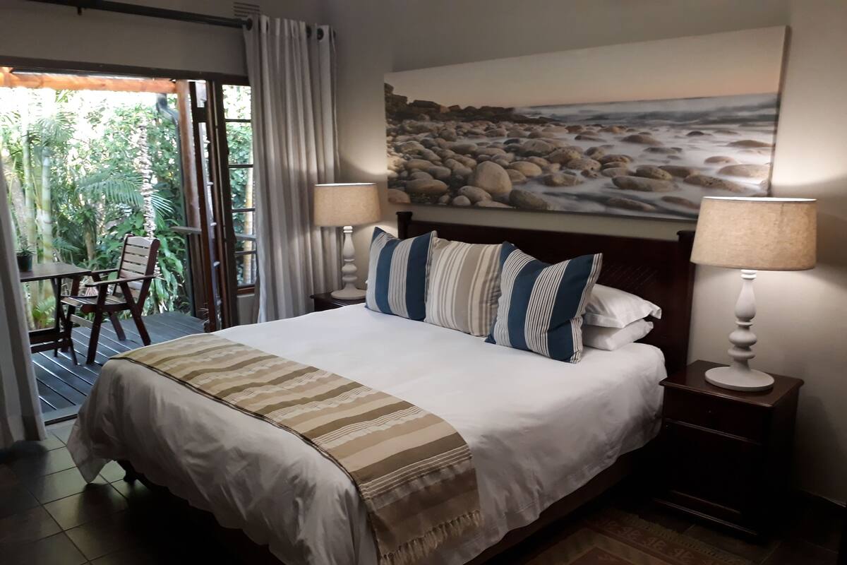 A hotel room with a large bed, a painting of a beach, a table with chairs, and a view of greenery through the window.