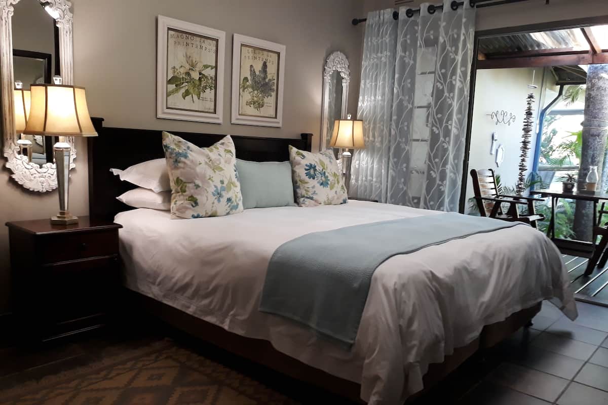 A neatly made bed with floral pillows, a nightstand with a lamp, and two framed botanical prints on the wall.