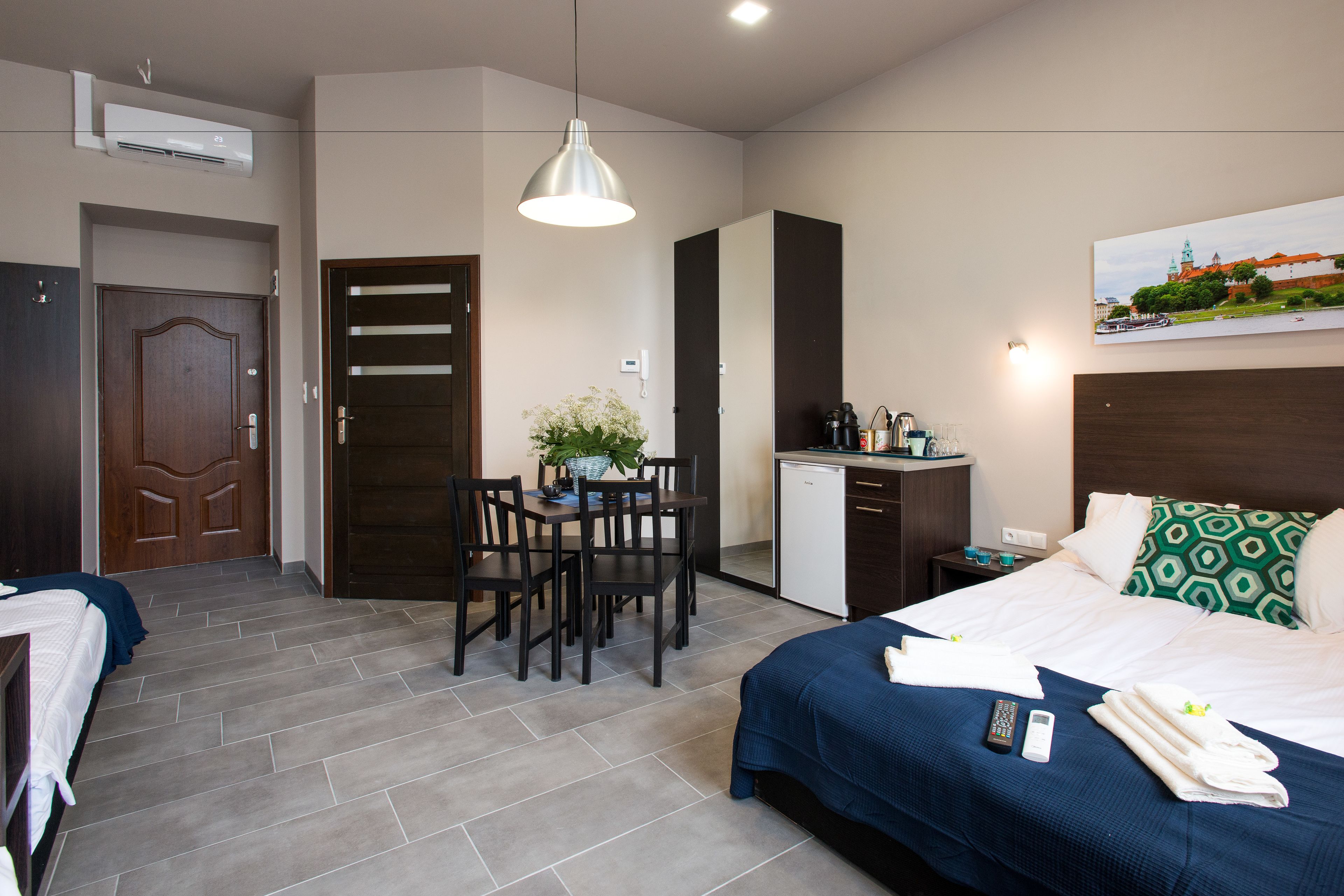 A hotel room with a bed, a desk, a chair, a small kitchenette, and a dining table.