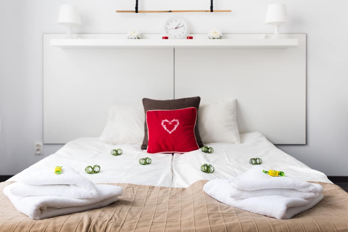 A neatly made bed with white linens, a brown headboard, and a red pillow with a heart design. There are two white lamps on a shelf above the bed.