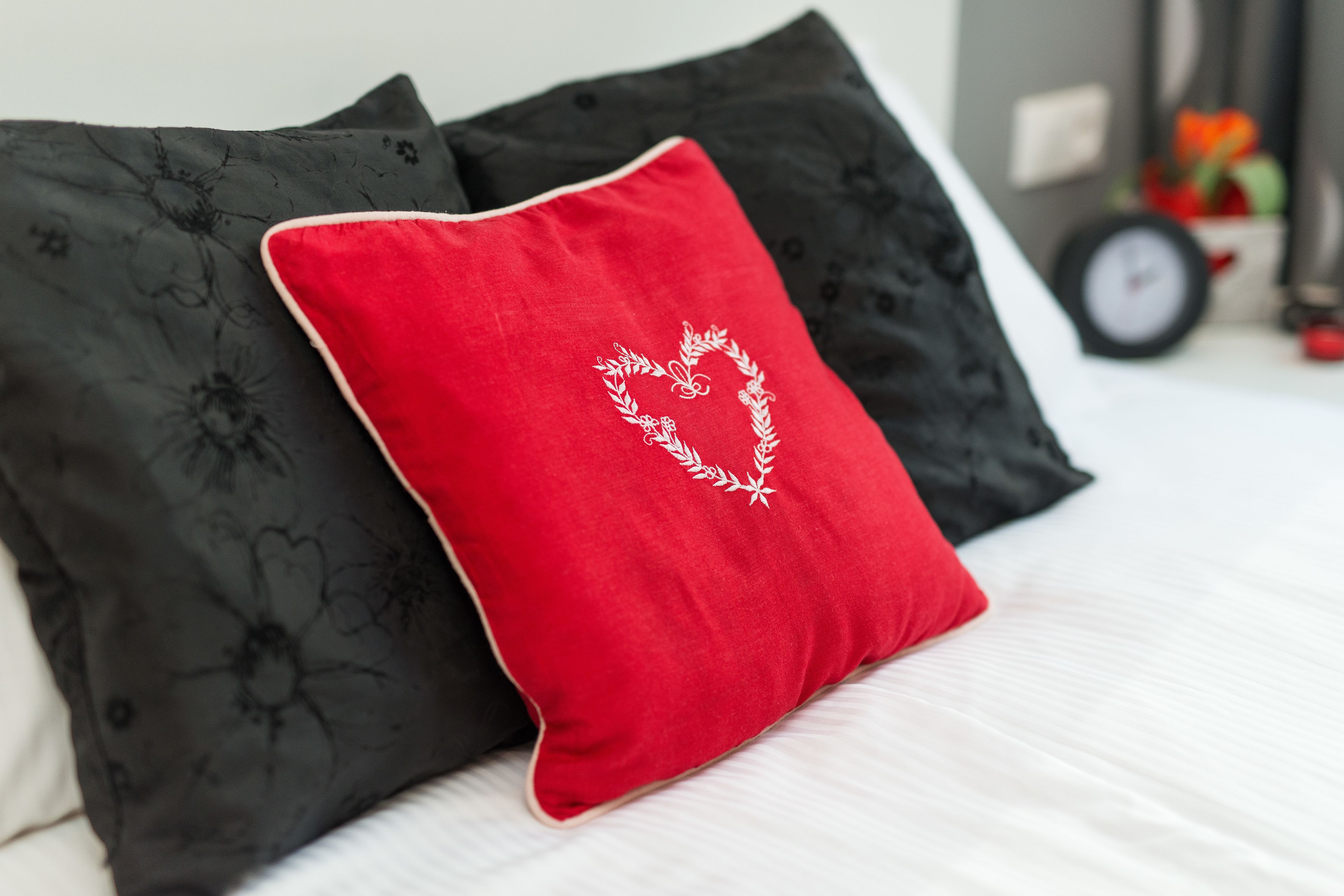 A bed with two pillows, one red and one black, with a clock and flowers in the background.