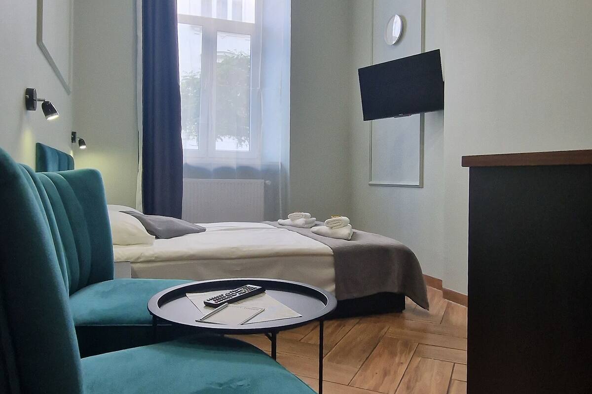 A modern hotel room with a teal sofa, a bed, a flat-screen TV, and a large window with curtains.