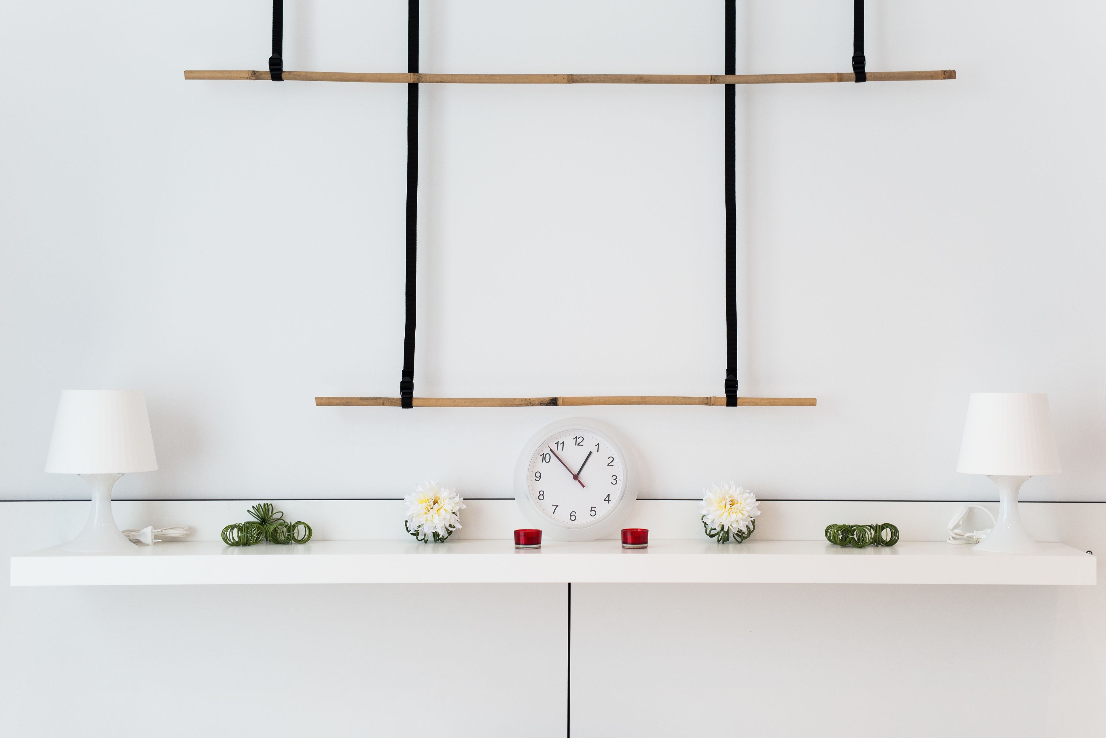 A minimalist room with a white wall, a wooden shelf, a white clock, two white lamps, and decorative flowers.