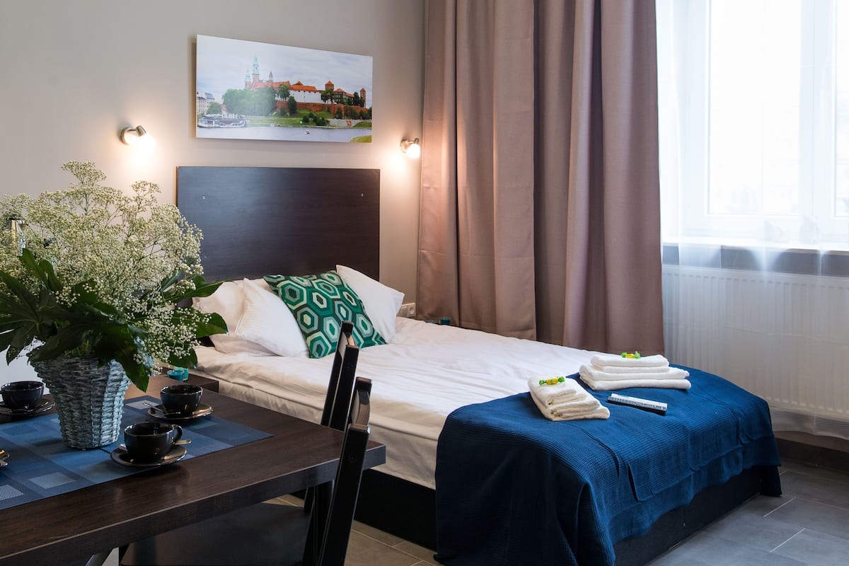A hotel room with a bed, a desk with a chair, a potted plant, and a painting on the wall.