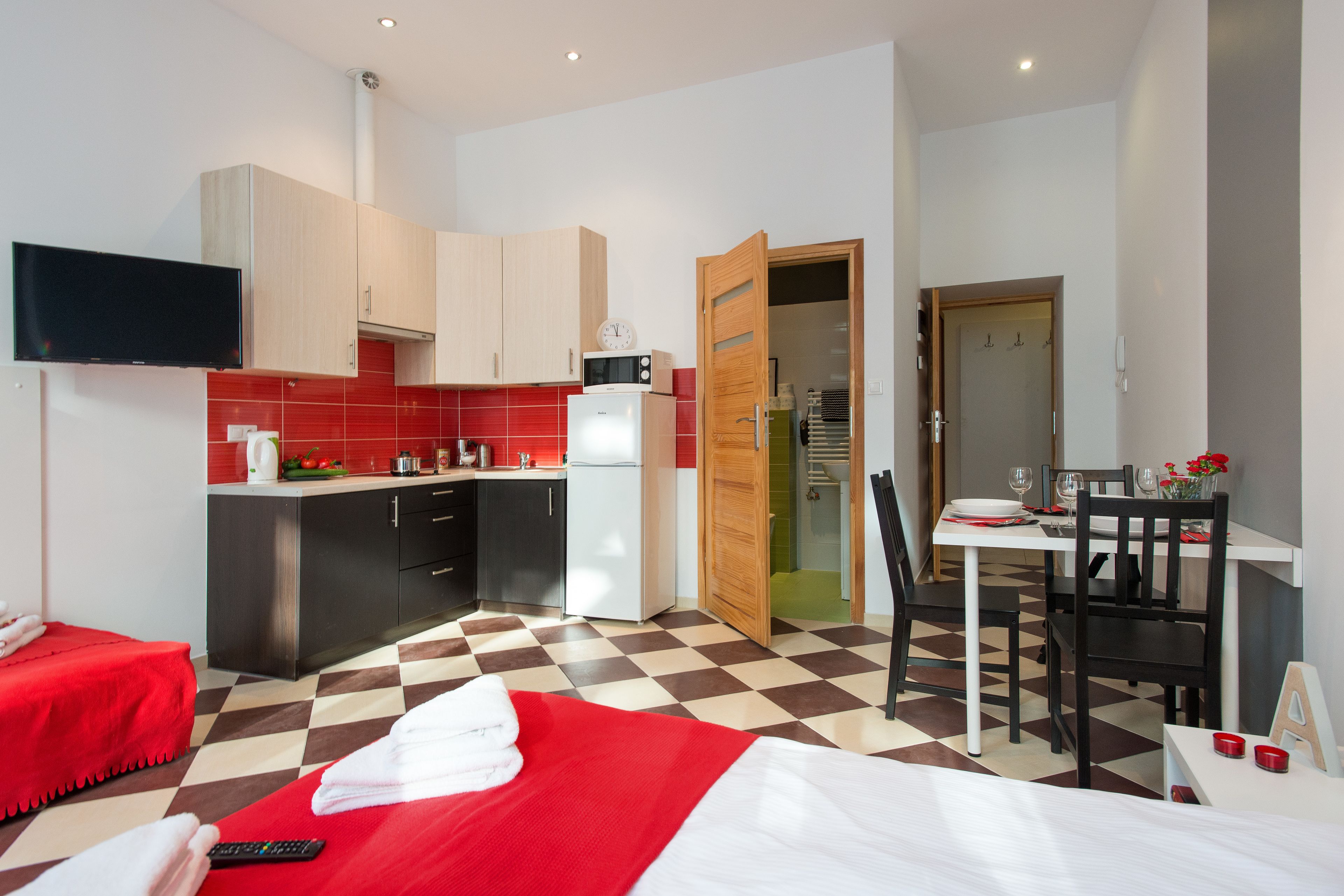 Una habitación de hotel moderna con piso a cuadros, una cama con ropa de cama roja, una pequeña cocina con microondas y refrigerador, un comedor con mesa y sillas, y un televisor montado en la pared.
