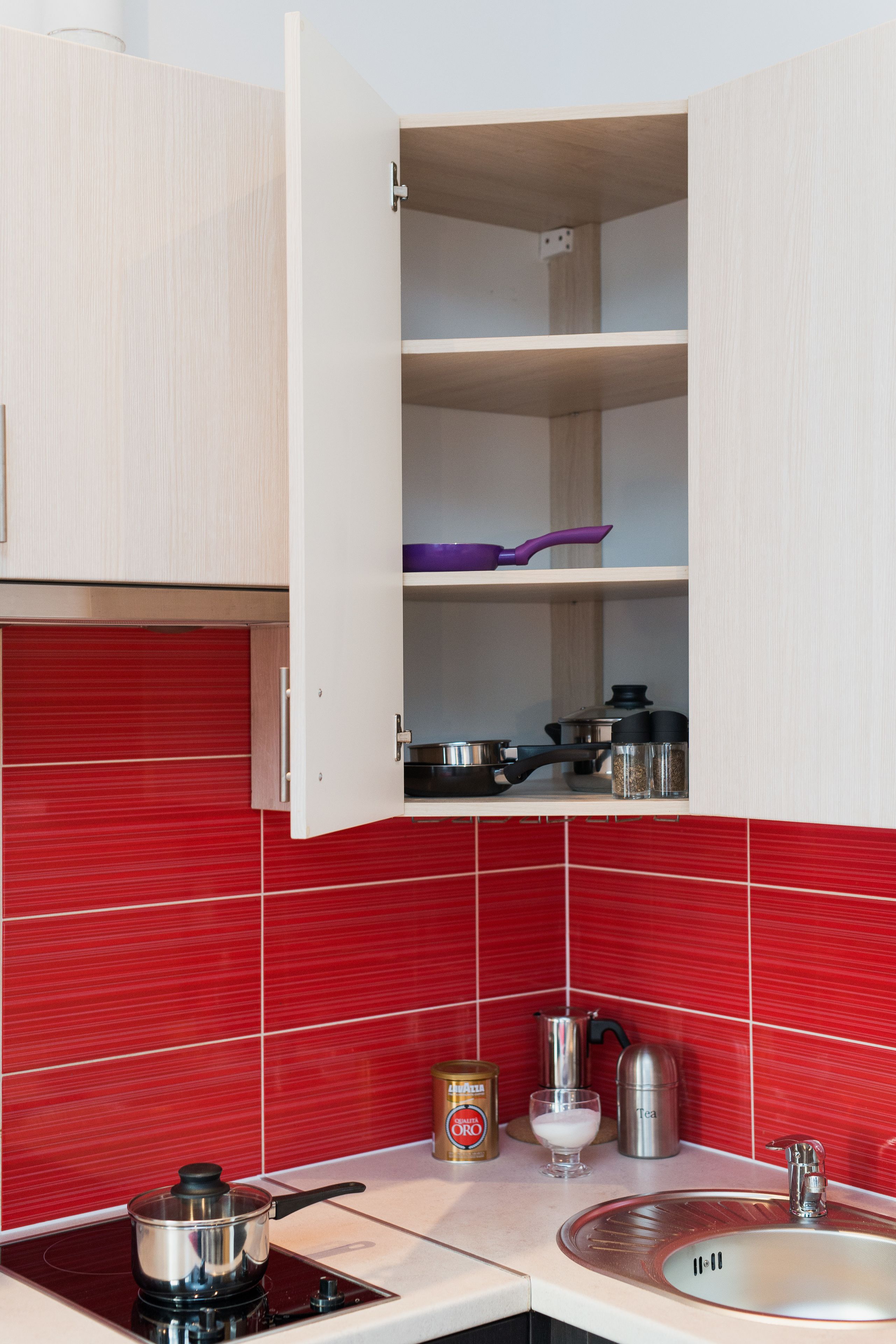 A kitchen with a red tiled backsplash, a white cabinet with open shelves, and a stainless steel kettle on the counter.