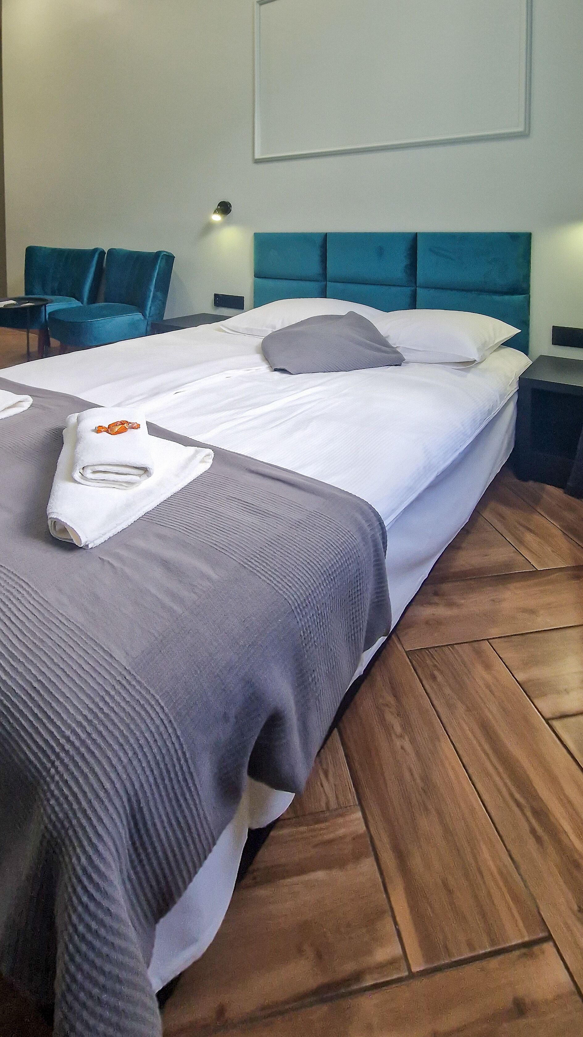 Un lit bien fait, avec une couverture grise et une serviette blanche enroulée, une tête de lit recouverte d’un tissu turquoise et un plancher en bois.
