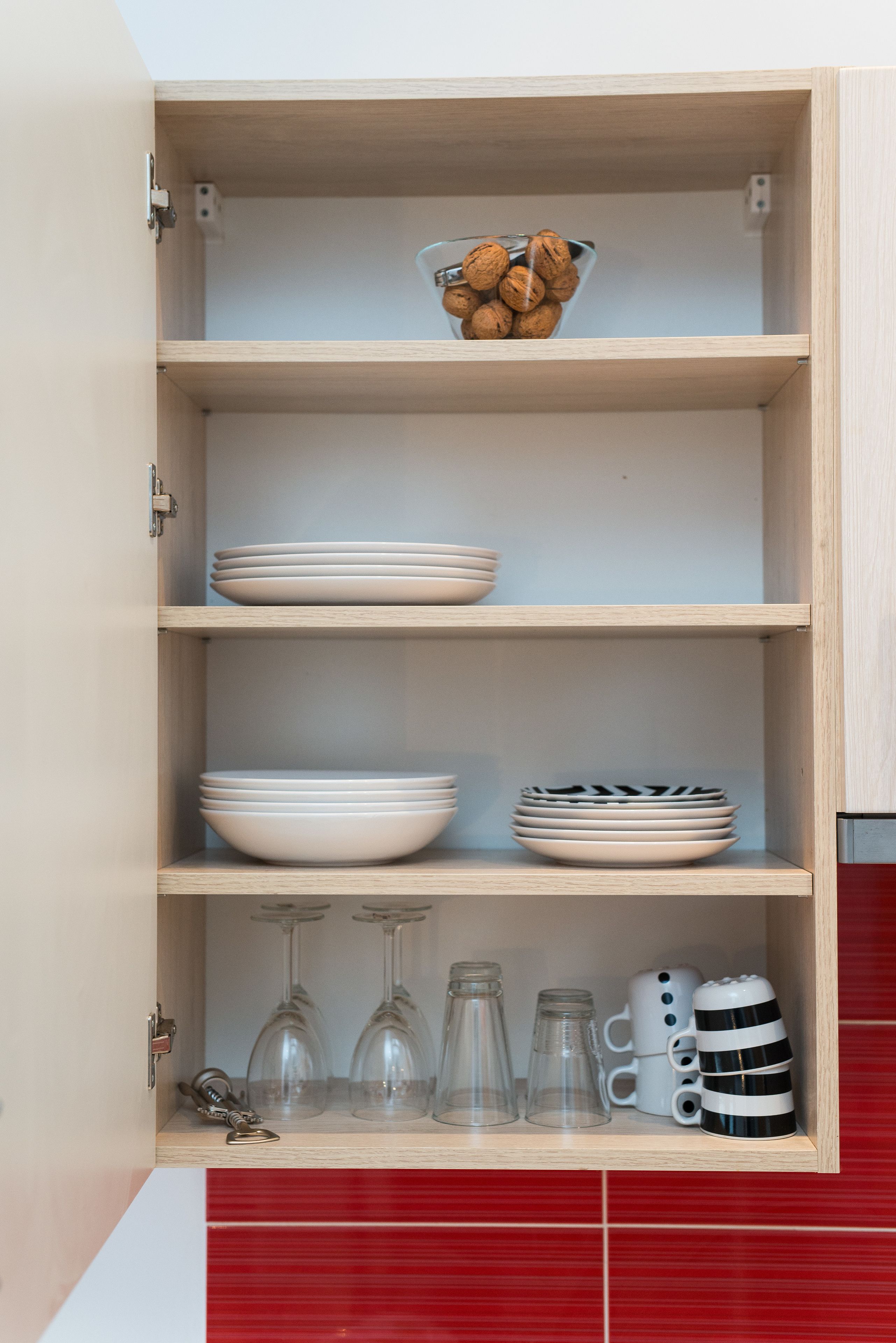 A kitchen cabinet with shelves holding white plates, bowls, a bowl of walnuts, wine glasses, and a striped mug.