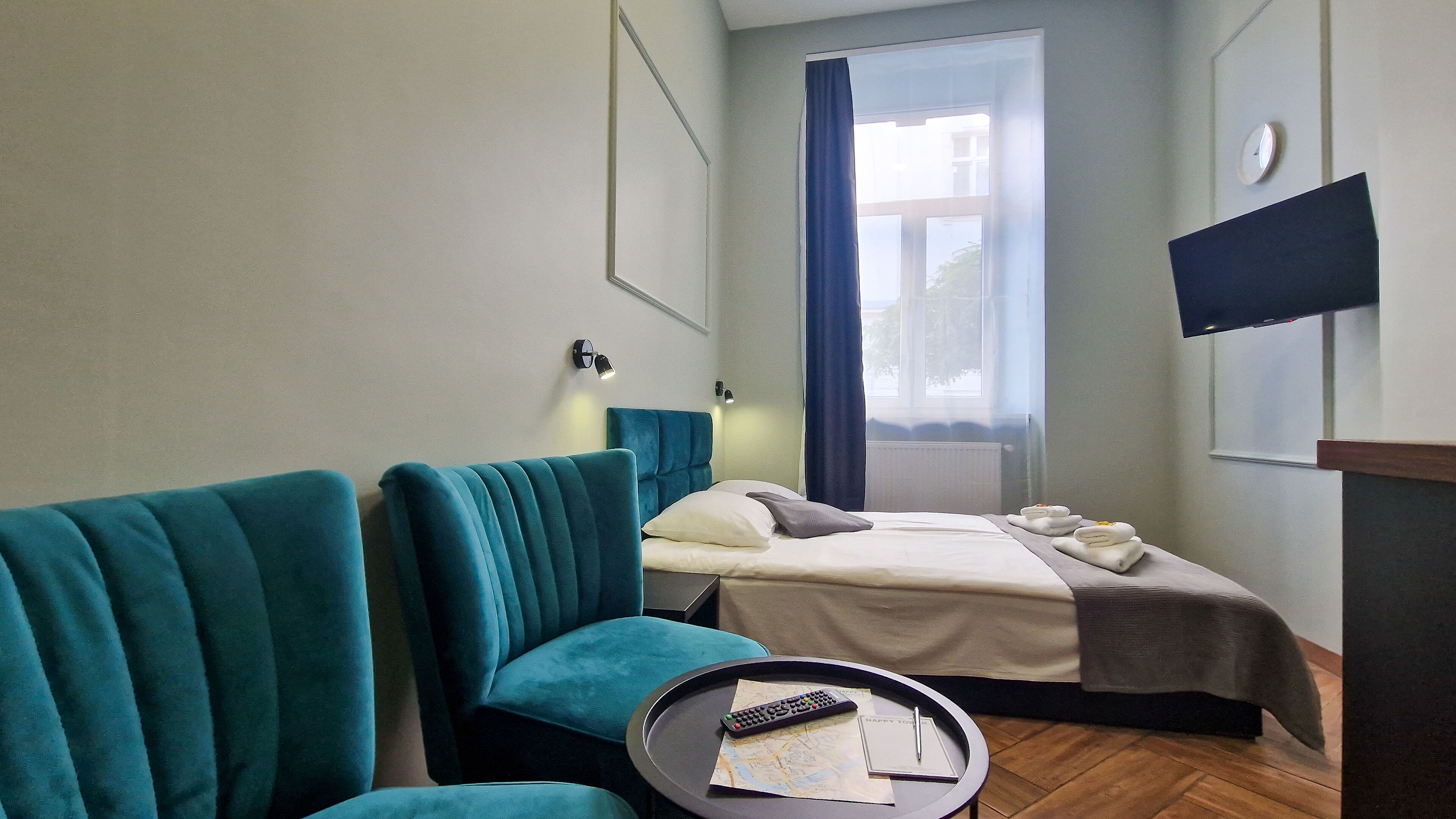 Quarto de hotel com uma cama, duas poltronas verde-água, uma mesinha de centro com um controle remoto e um mapa, e uma TV fixada na parede.