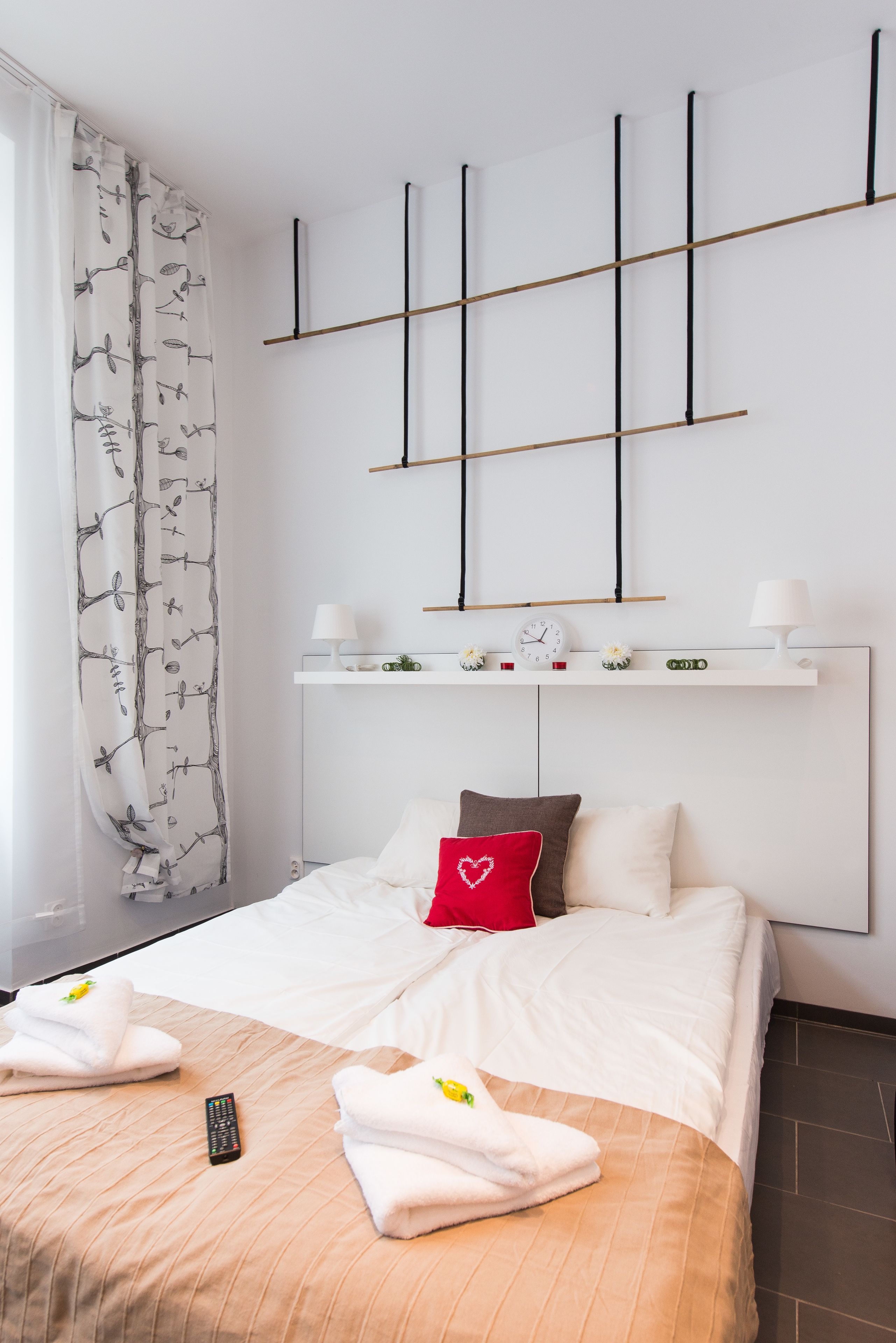 Habitación de hotel con una cama, un control remoto, toallas, un cojín con forma de corazón rojo y una estantería en la pared con relojes y flores.