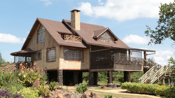 A large, rustic house with a prominent chimney, multiple windows, and a covered porch area. - Sangare Gardens (Mweiga)