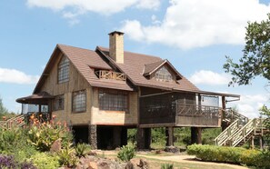 A large, rustic house with a prominent chimney, multiple windows, and a covered porch area. - Sangare Gardens (Mweiga)