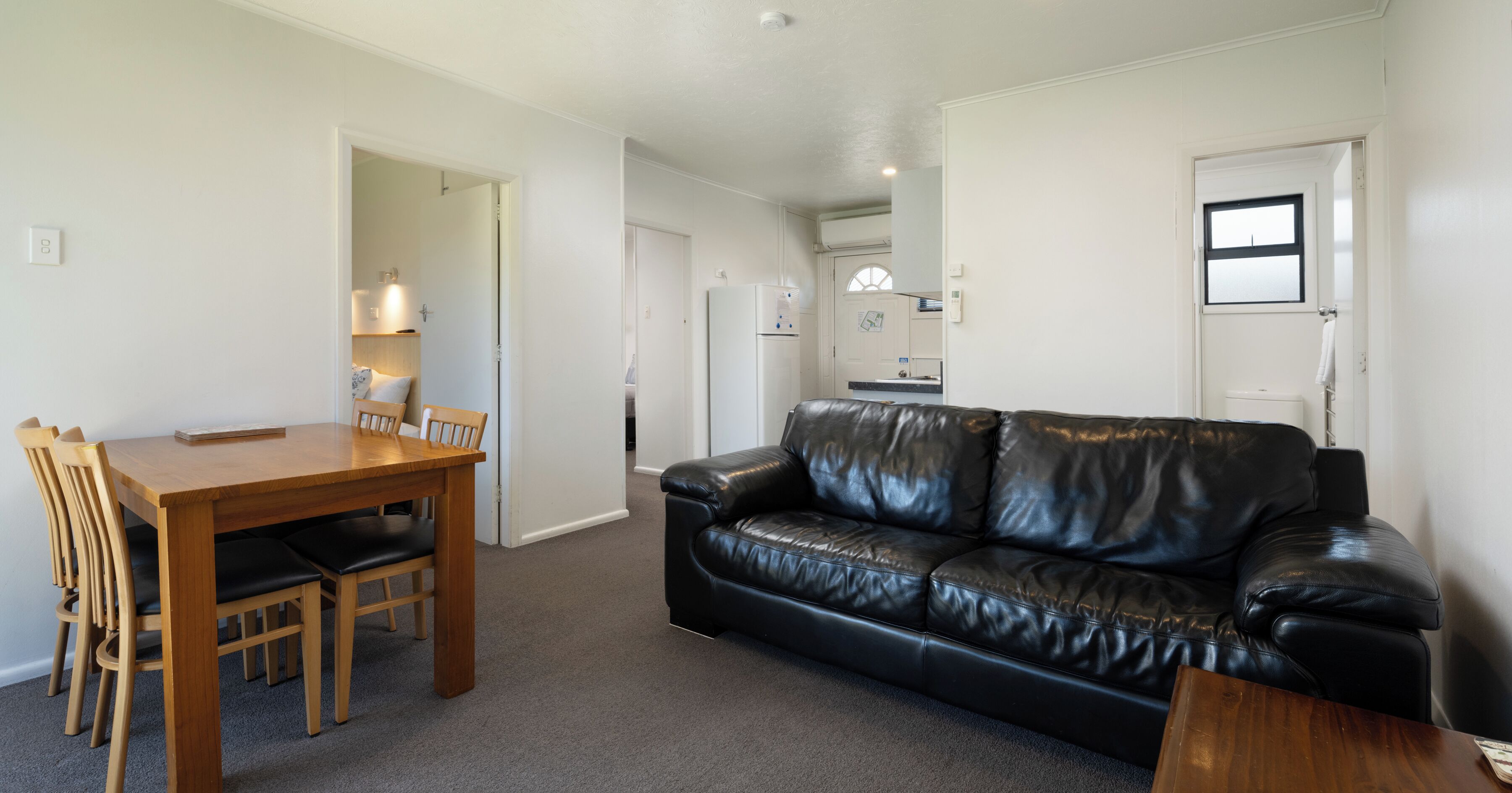 A living room with a wooden dining table, black leather sofa, and a kitchen area in the background.