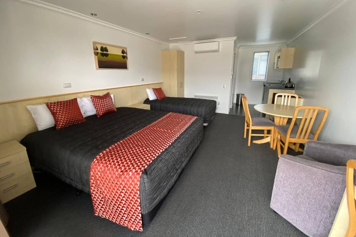 A hotel room with two beds, a dining table, chairs, a small kitchenette, and a painting on the wall.