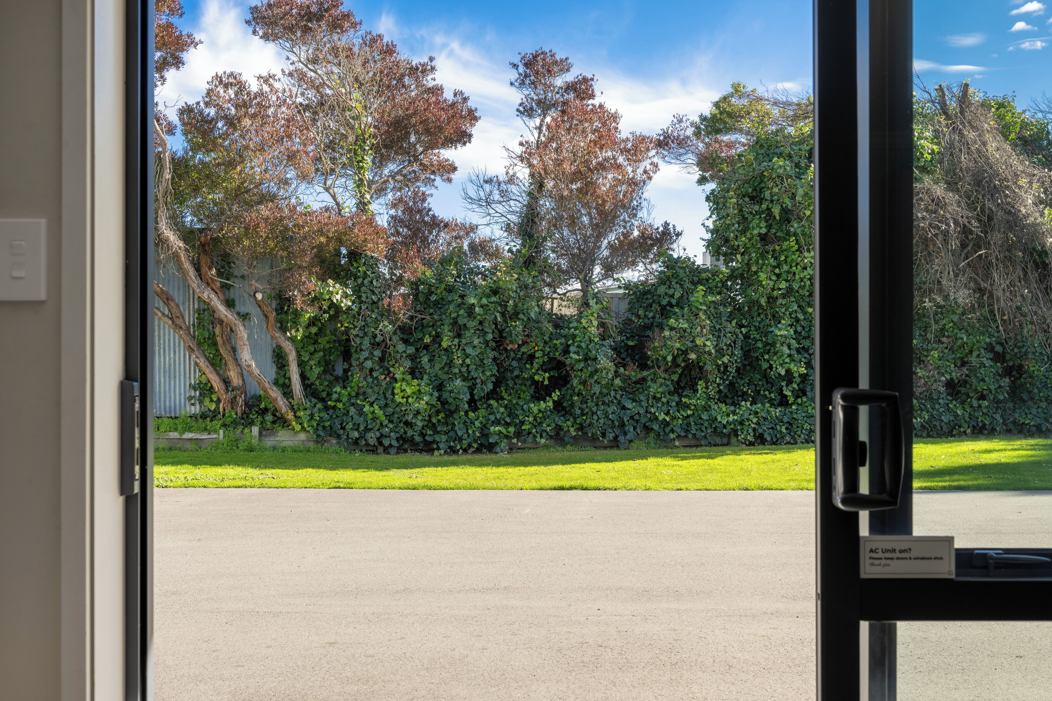 A view through a glass door of a parking area with a green lawn and trees.
