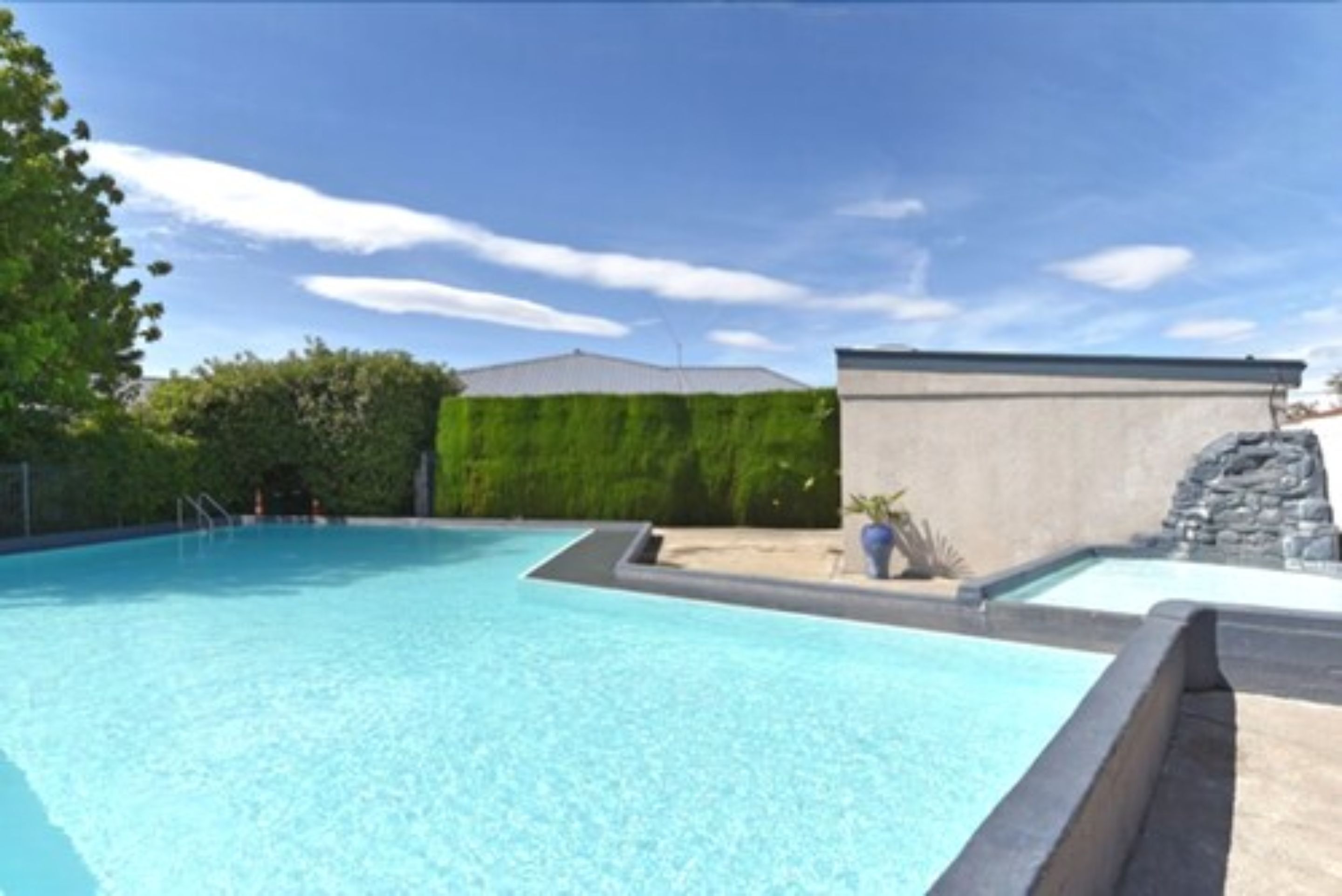 A swimming pool with a clear blue water, surrounded by a stone wall and a hedge.
