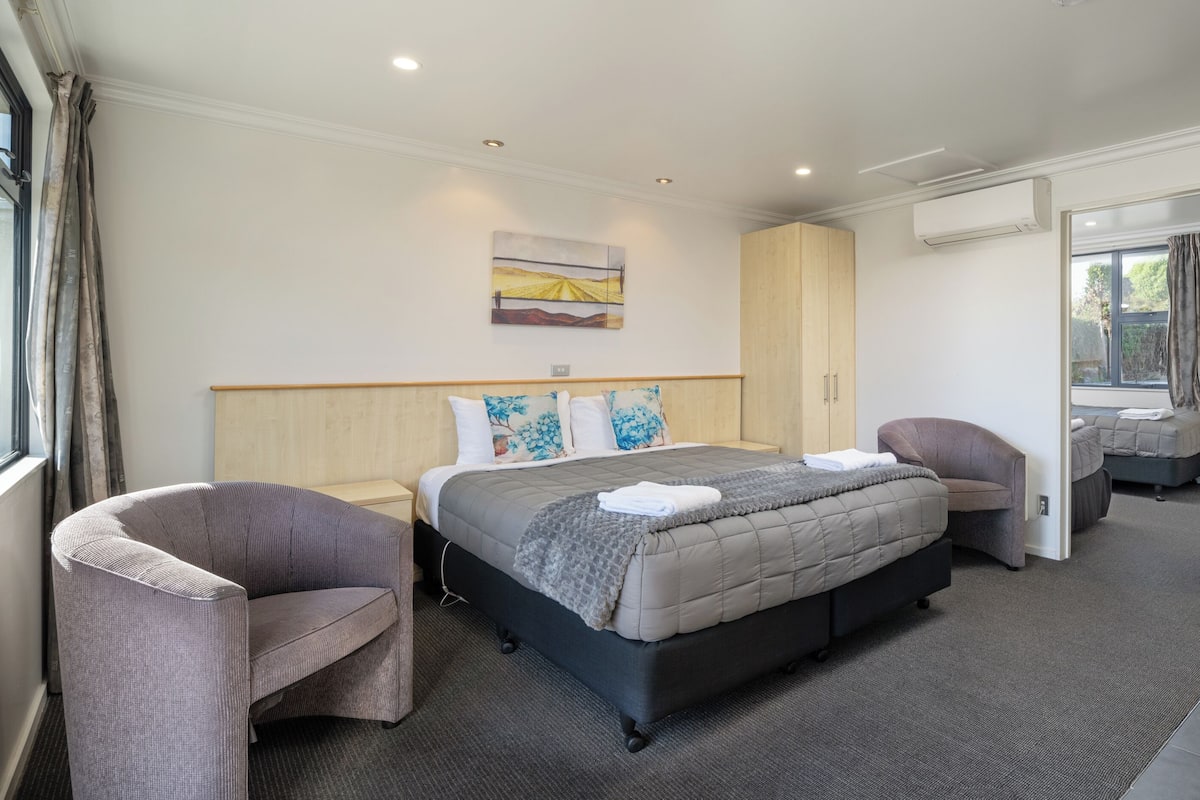 A modern hotel room with a large bed, two armchairs, a small table, and a view of the outdoors through a sliding door.