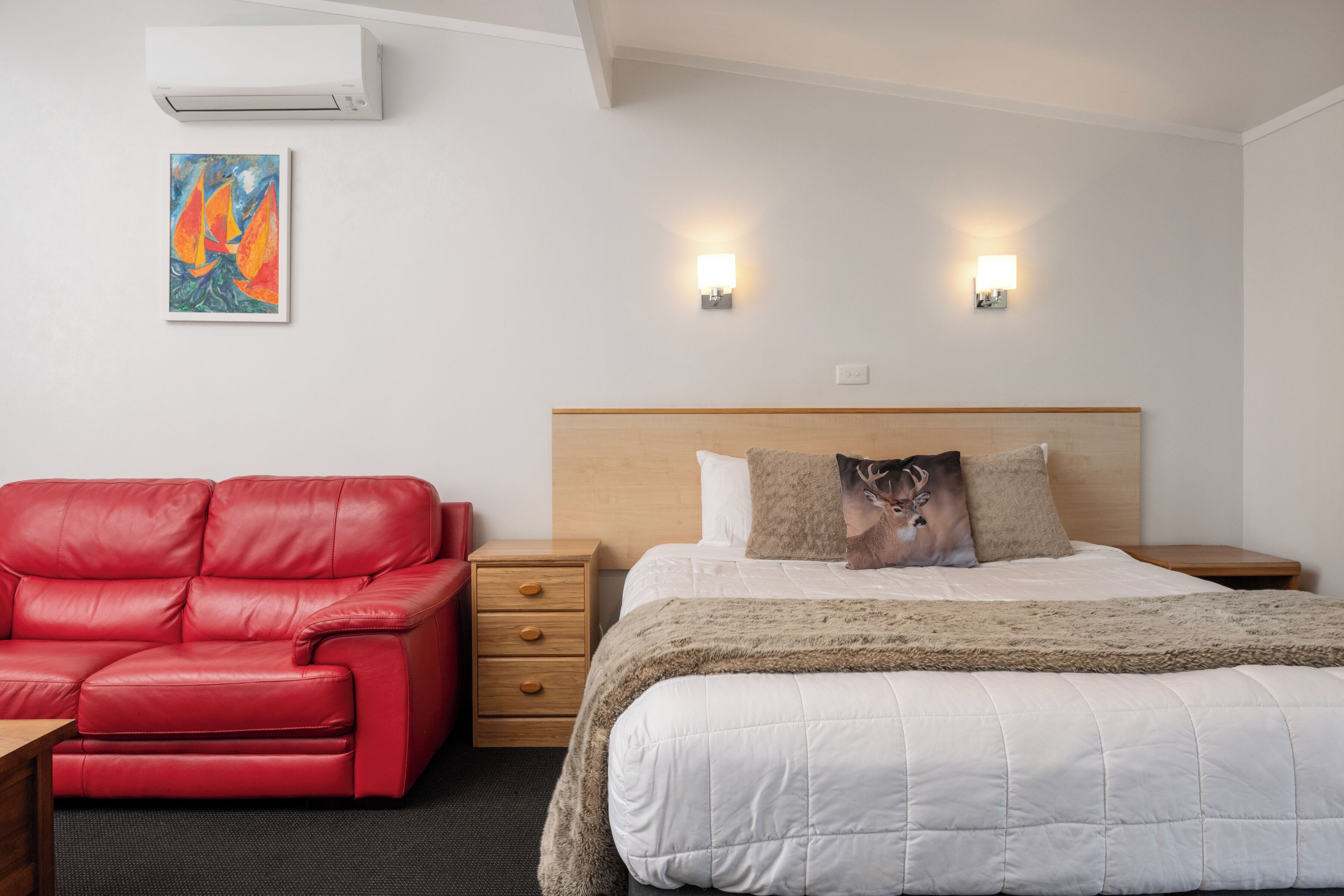 A hotel room with a bed, a red leather sofa, a wooden nightstand, and a painting on the wall.