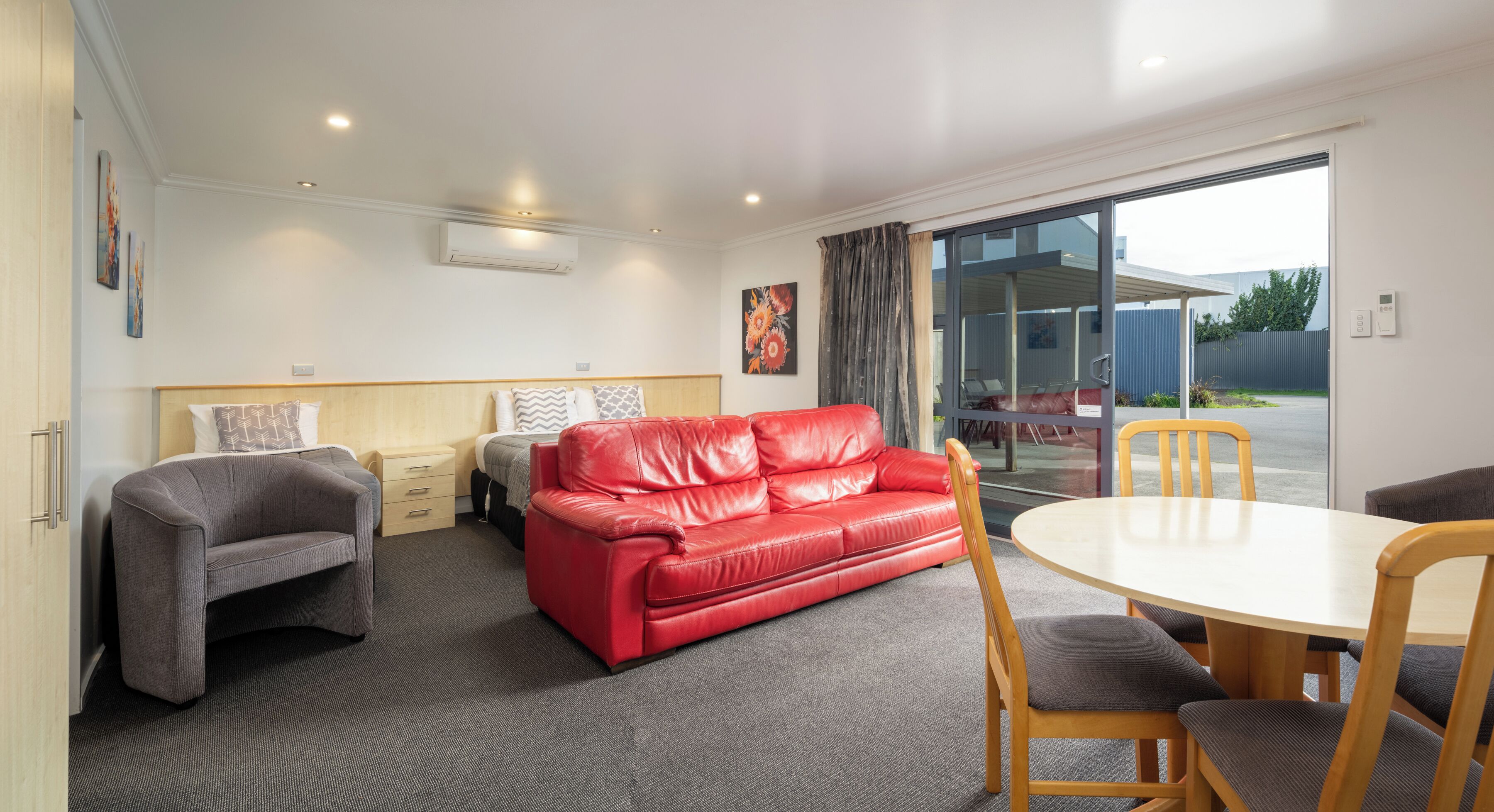 A modern hotel room with a red leather sofa, a grey armchair, a wooden dining table, and a large window offering a view of the outside.