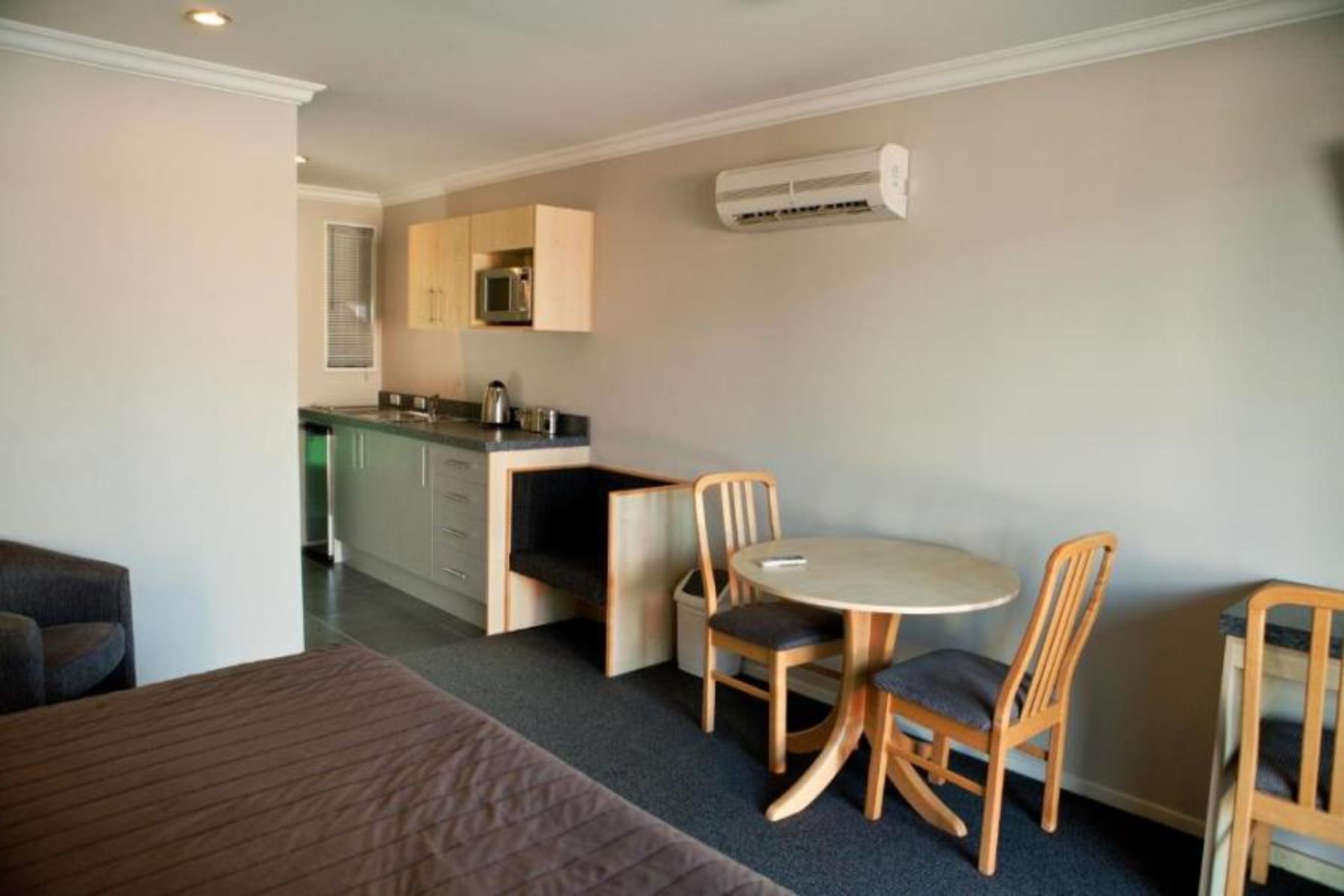 A compact room with a bed, a small dining table with chairs, a kitchenette, and an air conditioning unit.