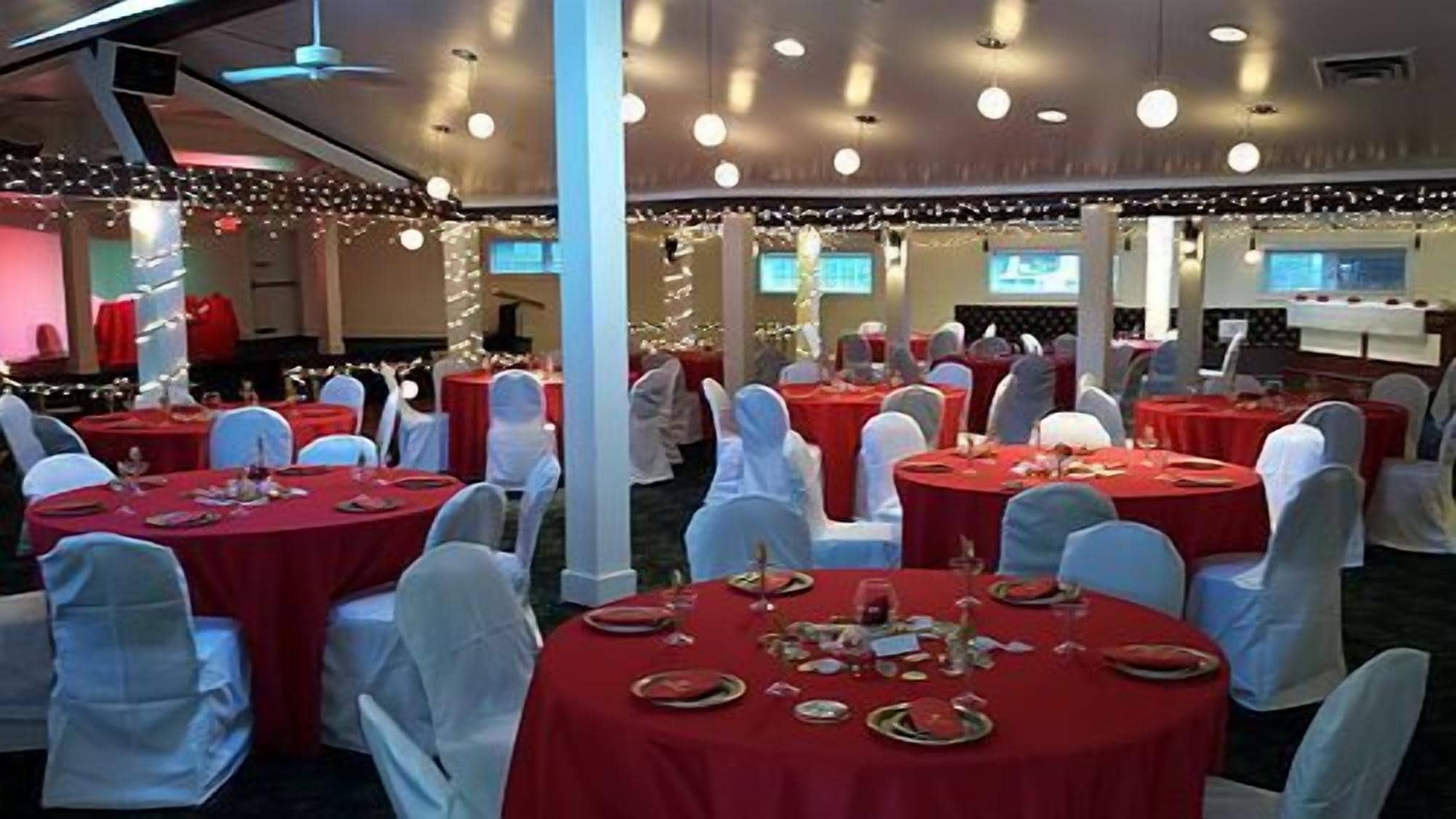 Un salón de banquetes con mesas redondas cubiertas con manteles rojos, fundas blancas para las sillas y con platos, vasos y cubiertos.