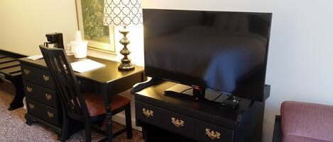 A room with a black TV stand, a desk, a chair, and a lamp.