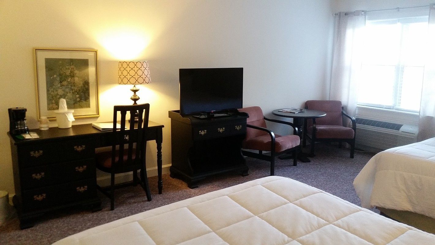 A hotel room with a desk, a television, a chair, a small table, and two beds.
