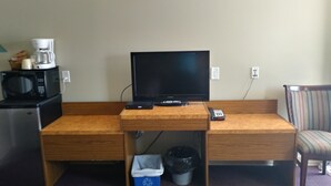 Habitación de hotel compacta con escritorio de madera, televisor, microondas, cafetera y basurero.