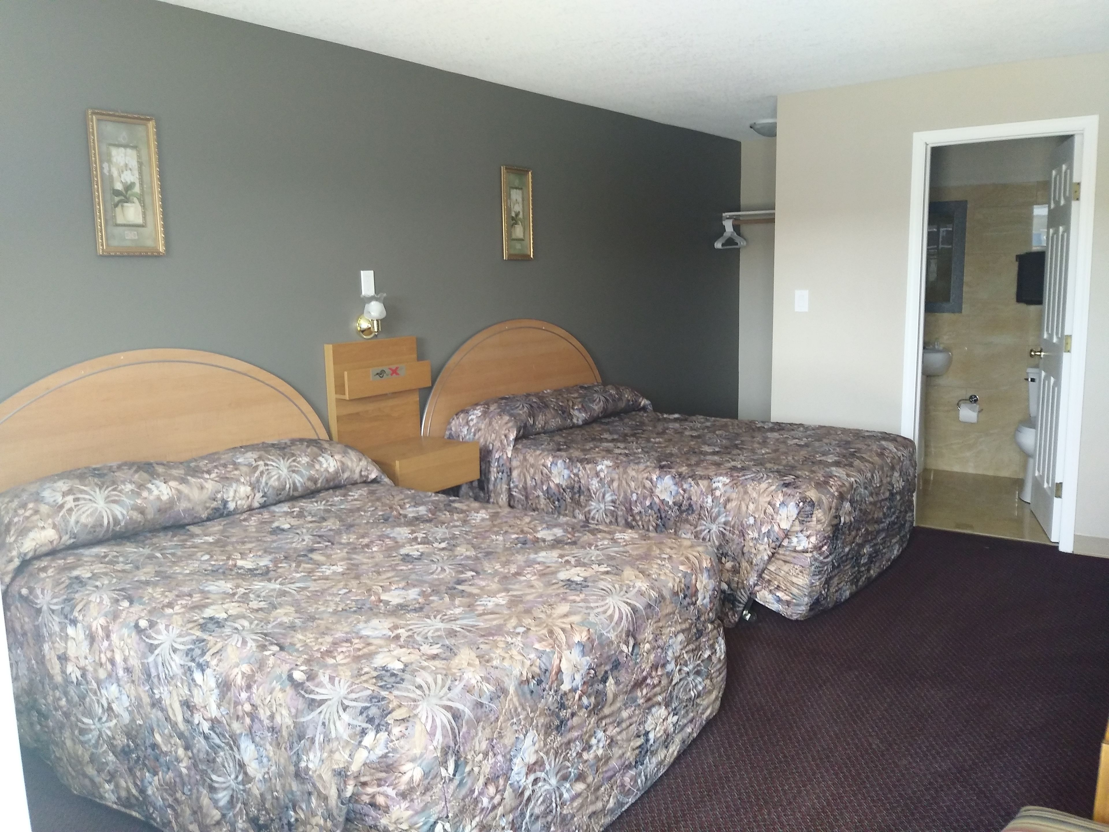 A hotel room with two beds, a wooden nightstand, a wall-mounted lamp, and a bathroom visible through an open door.