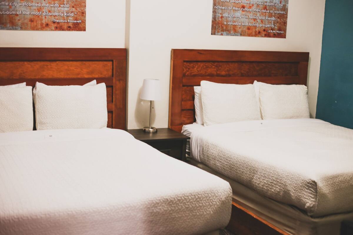 A hotel room with two beds, each with white linens and wooden headboards. There is a nightstand with a lamp between the beds.