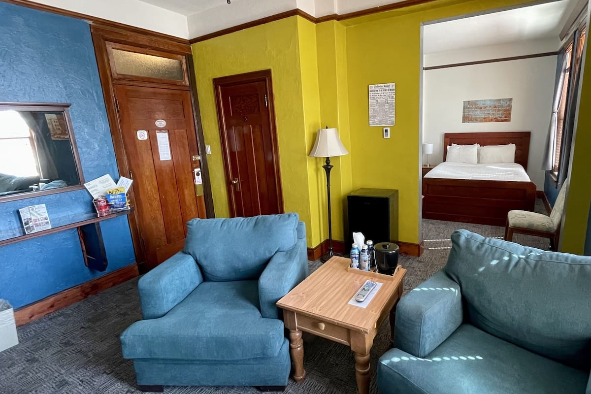 A hotel room with a bed, two blue armchairs, a wooden table, a lamp, a window with blinds, and a door.