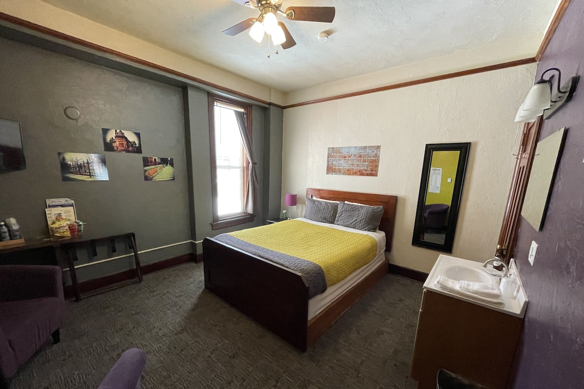 A hotel room with a bed, a desk, a chair, a window with curtains, a ceiling fan, and a mirror.