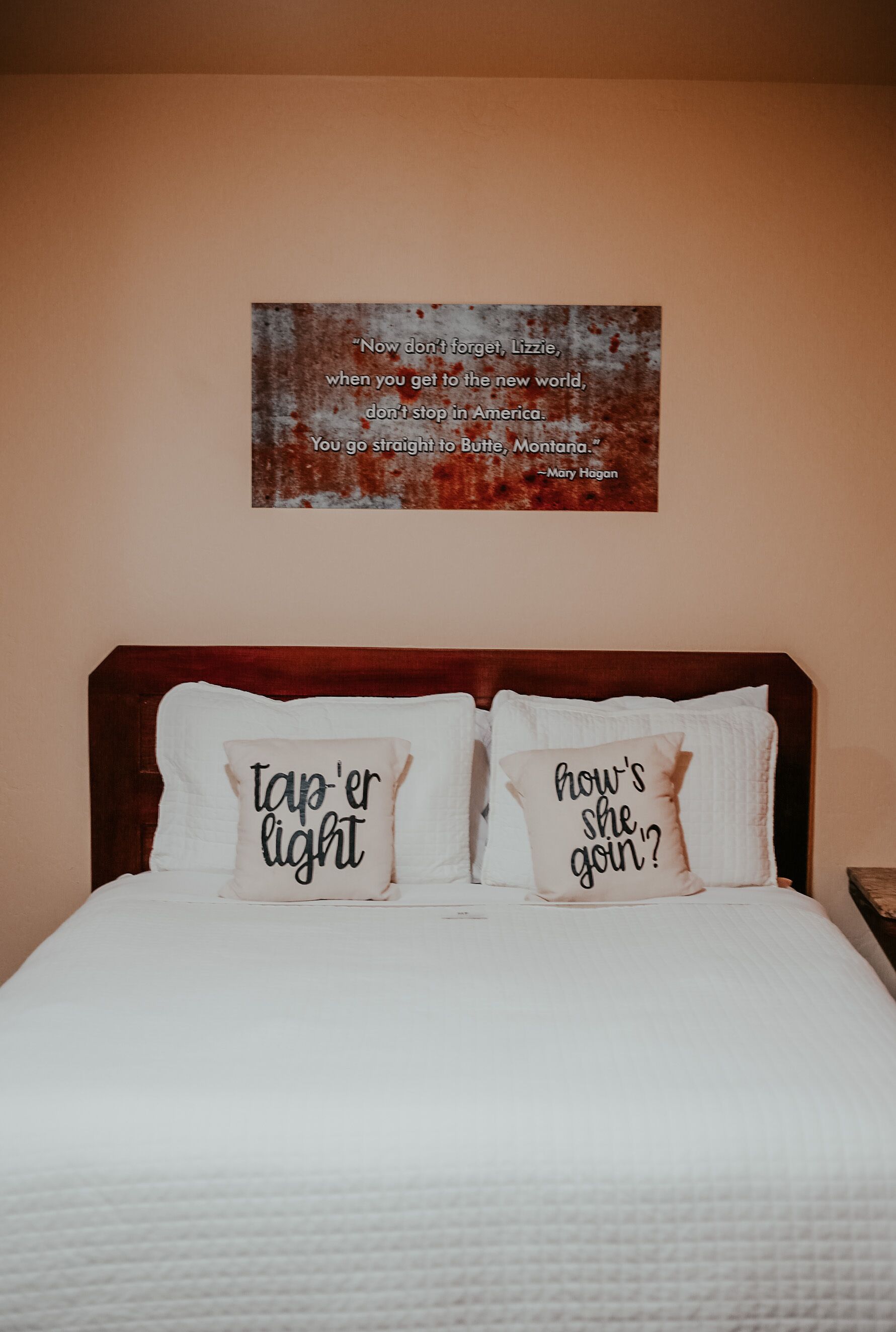 Una cama bien hecha con dos almohadas, una con la frase 'tap' er light' y la otra con 'how's she goin'?'. 