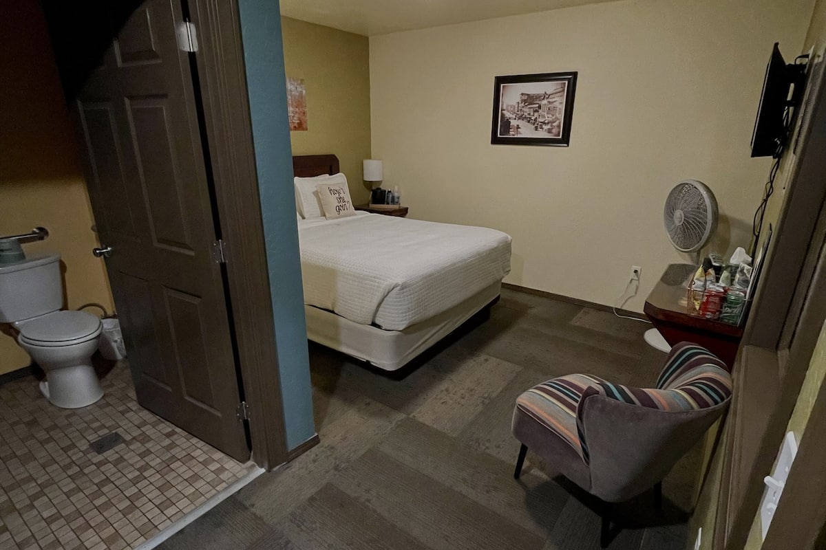 A bedroom with a bed, a chair, a bedside table with a lamp, a fan, and a framed picture on the wall.