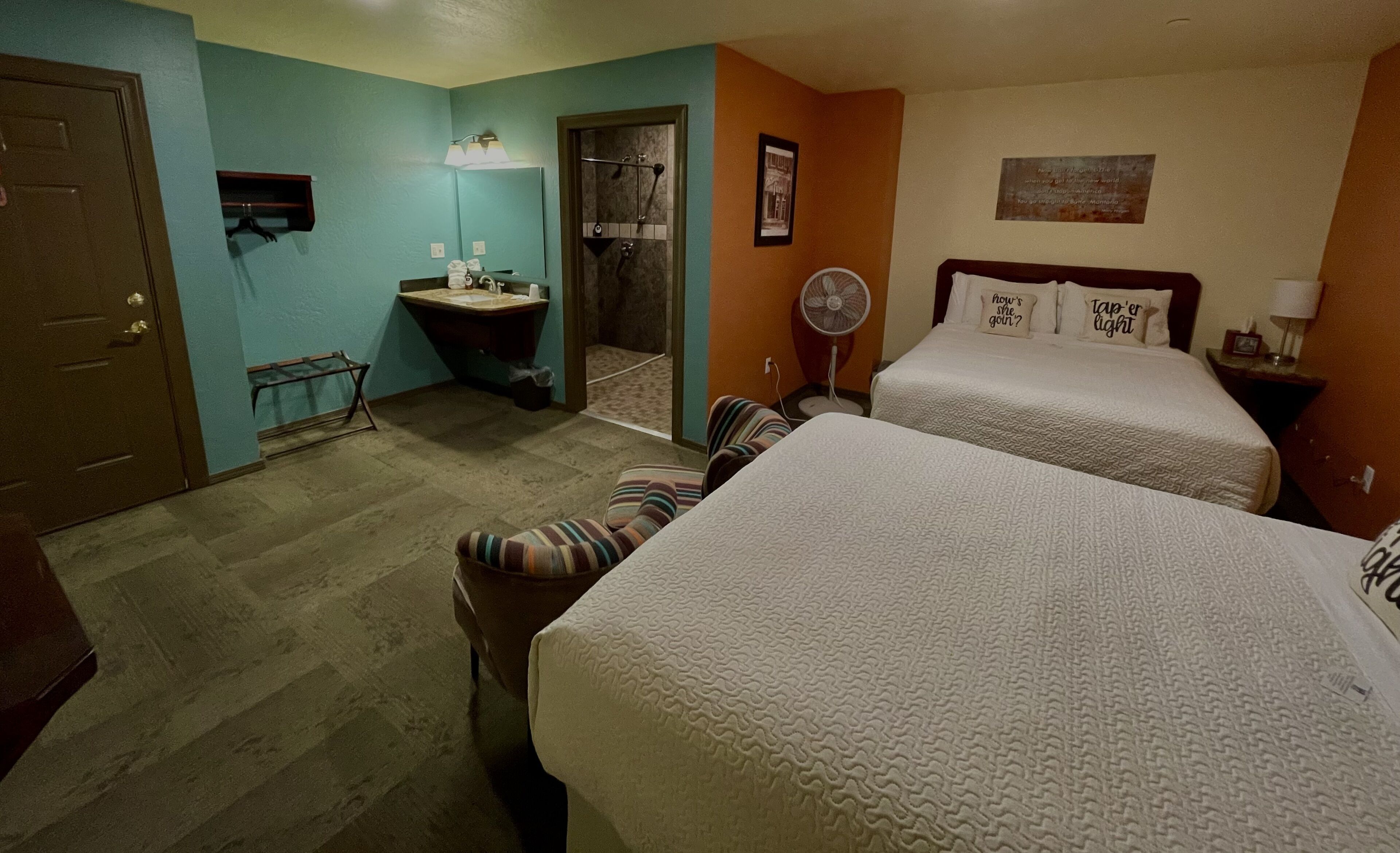A hotel room with two beds, a bathroom with a sink and mirror, a fan, and a door.