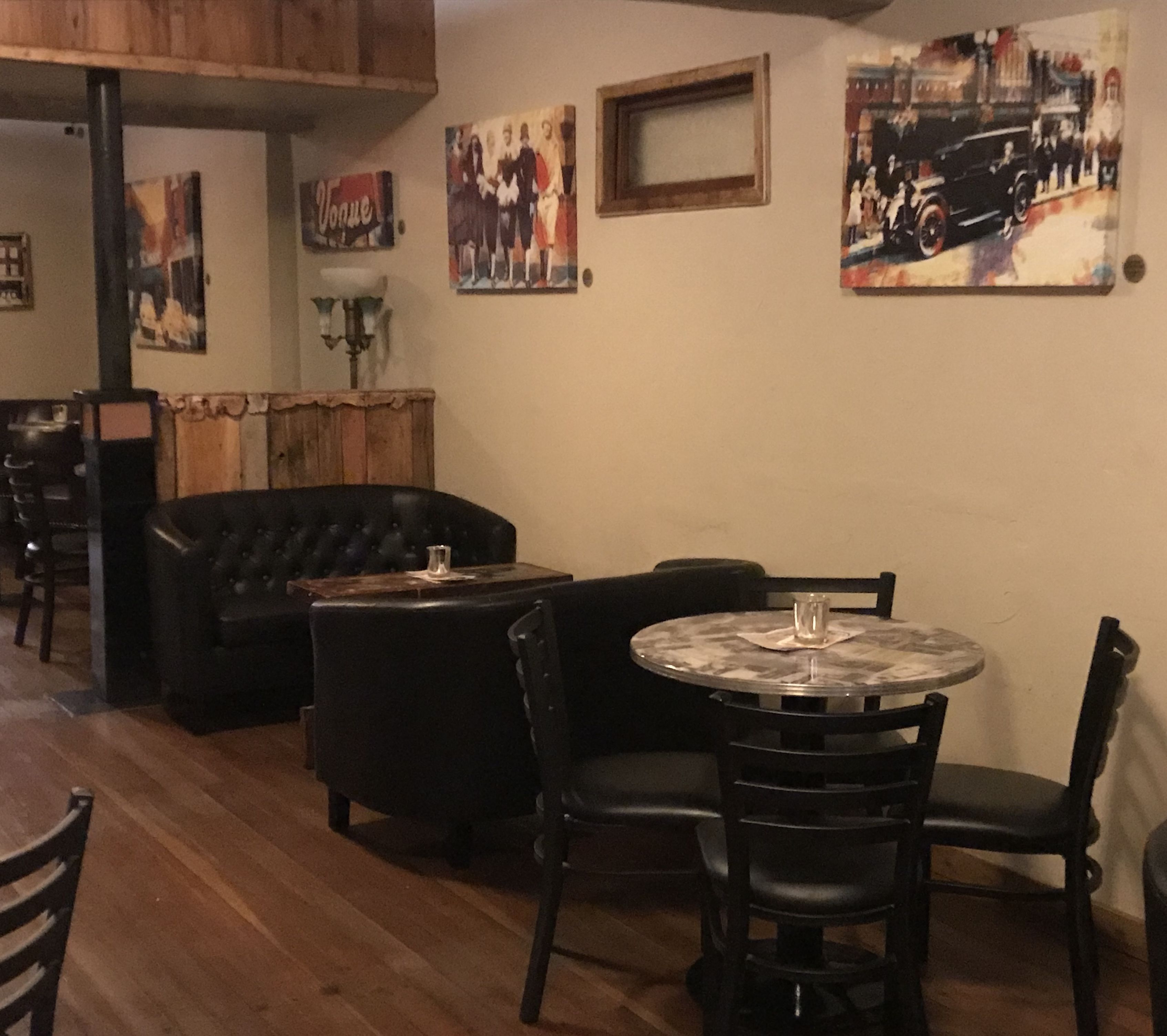 A cozy restaurant with a wooden floor, black leather seating, and a round table with chairs. There are wall-mounted paintings and a framed picture above the table.