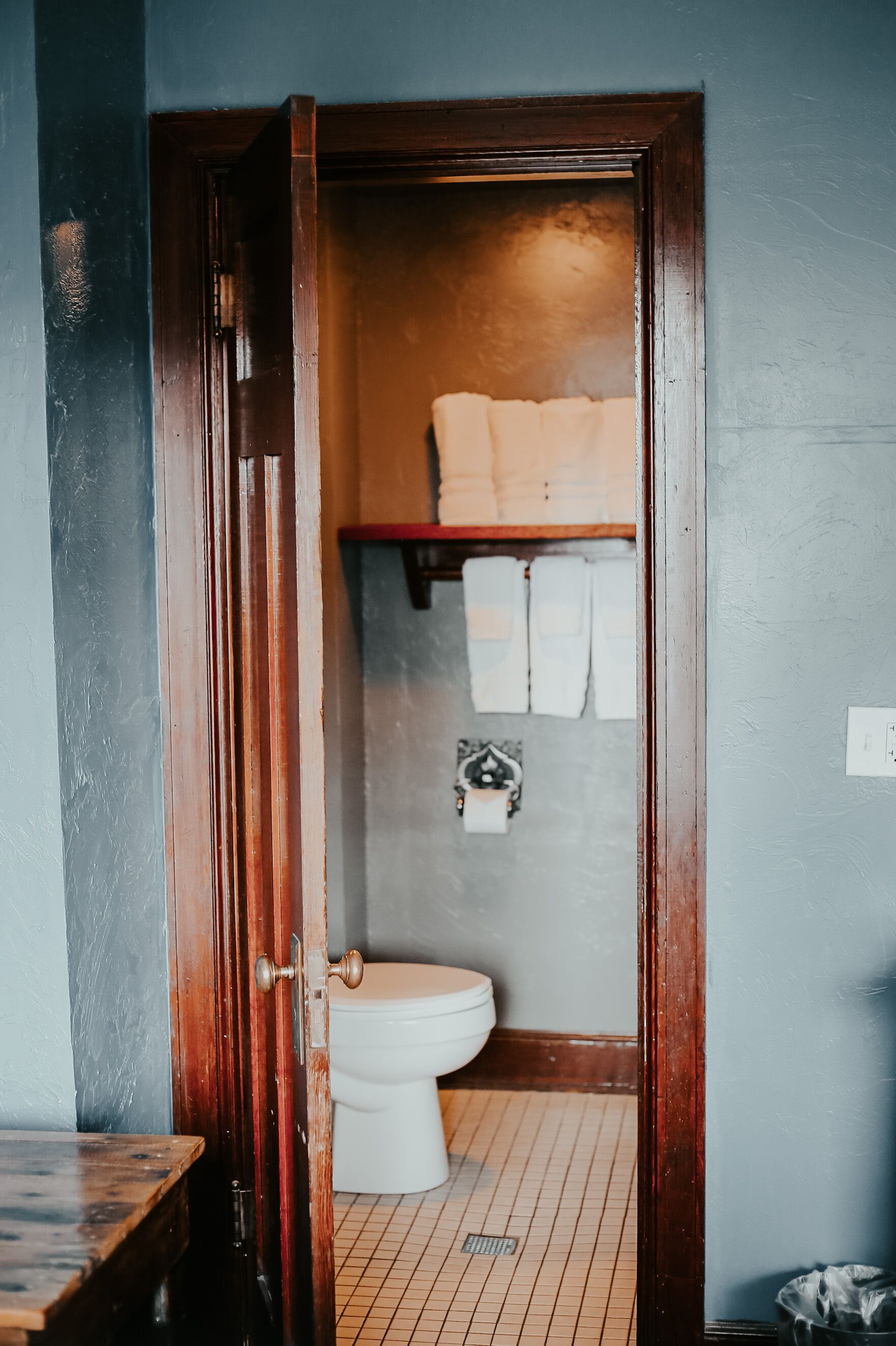 A wooden door leading to a bathroom with a toilet, a shelf with towels, and a tiled floor.